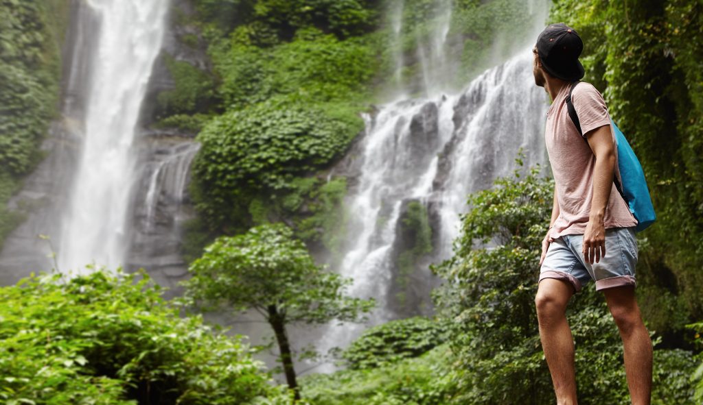 Full-length portrait of young hiker or adventurer in denim shorts and snapback enjoying nature, standing barefooted on rocks and looking back at gorgeous waterfall during his trekking trip in woods